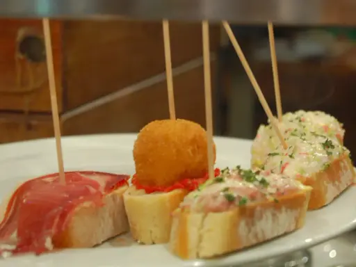 Różnorodne pinchos na białym talerzu: z szynką, kulką w panierce, pastą rybną i sałatką.