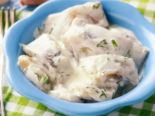 Śledzie w śmietanie, jak u babci. Delikatne kawałki ryby w kremowym sosie ze szczypiorkiem, podane w niebieskiej miseczce.