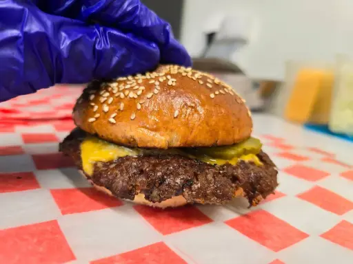 Rękawiczka w niebieskiej rękawiczce przygotowuje pysznego smash burgera z serem i ogórkiem.