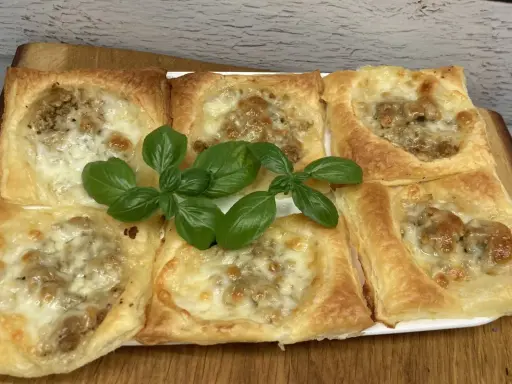 Sześć kwadratowych porcji ciasta francuskiego z mięsem mielonym i serem, ozdobione listkami bazylii.