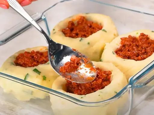Łyżka nakłada mięsne nadzienie do wgłębień w ziemniaczanych grule, przygotowywanych w szklanym naczyniu.