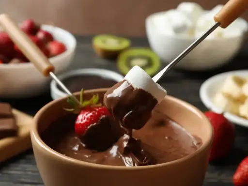 Pyszne fondue czekoladowe z pianką i truskawką na patyczku. W tle owoce i pianki.