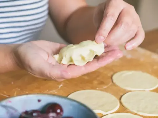 Dłonie formują pieroga z ciasta na pierogi bez jajka. Na blacie leżą okrągłe placki ciasta i miska z wiśniami.