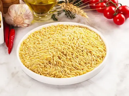 Biała miska pełna makaronu w kształcie ryżu, obok czosnek, chili, oliwa i pomidorki.