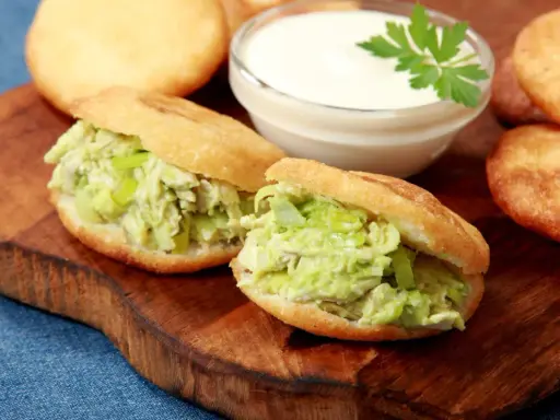 Dwie pyszne arepas z kurczakiem i awokado, obok miseczka sosu i kilka niefaszerowanych arepas.