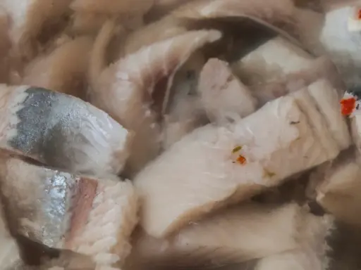 Kawałki kiszonych śledzi w zalewie, z widocznymi fragmentami skórki i przyprawami.