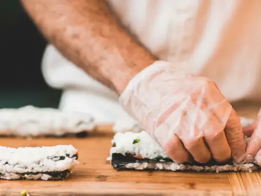 Ręce w rękawiczkach zwijają sushi. Na desce leżą gotowe rolki i składniki do kolejnej. Idealna zalewa do sushi podkreśli smak.