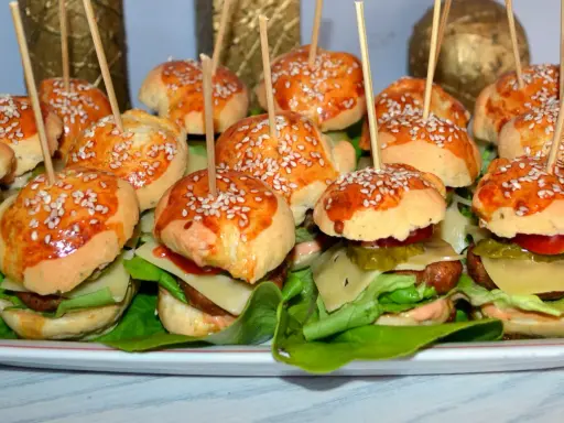 Mini hamburgery z sezamem na patyczkach, ułożone na talerzu z sałatą i dodatkami.