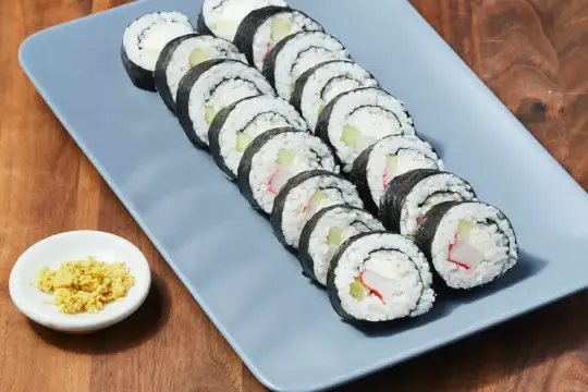 Rząd roladek sushi z kremowym serkiem, ogórkiem i paluszkami krabowymi na szarym talerzu. Obok mała miseczka z imbirem.