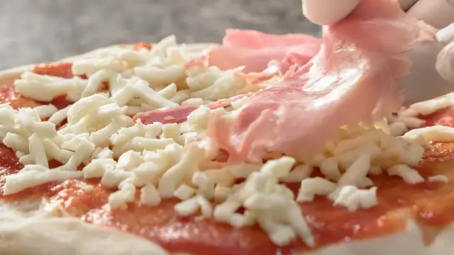Rękawiczki zakładają cienkie plastry szynki parmeńskiej na pizzę z serem i sosem pomidorowym.