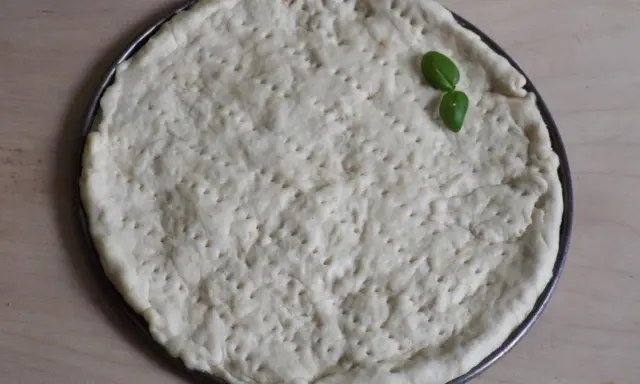 Gotowy do pieczenia, okrągły spód do pizzy z dziurkami, ozdobiony listkiem bazylii. Idealny szybki przepis na ciasto do pizzy.