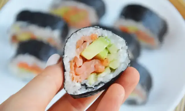 Ręka trzyma kawałek futomaka z łososiem i awokado. Na talerzu widać więcej sushi.