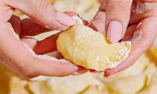 Dłonie z różowymi paznokciami lepią pierogi. Mąka lekko oprósza palce i ciasto. To idealny przepis na domowe pierogi.