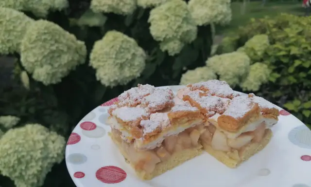 Pyszna szarlotka z bezą i kruszonką, oprószona cukrem pudrem, na talerzu wśród kwitnących hortensji.