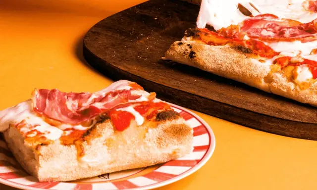 Kawałek pysznej pizzy rzymskiej z szynką i ciągnącym się serem na talerzu, obok reszta na desce.