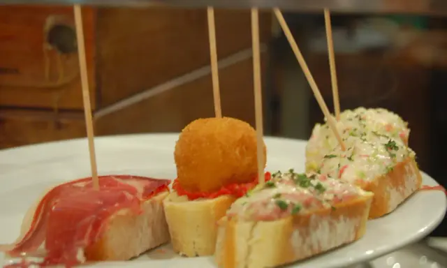 Różnorodne pinchos na białym talerzu: z szynką, kulką w panierce, pastą rybną i sałatką.
