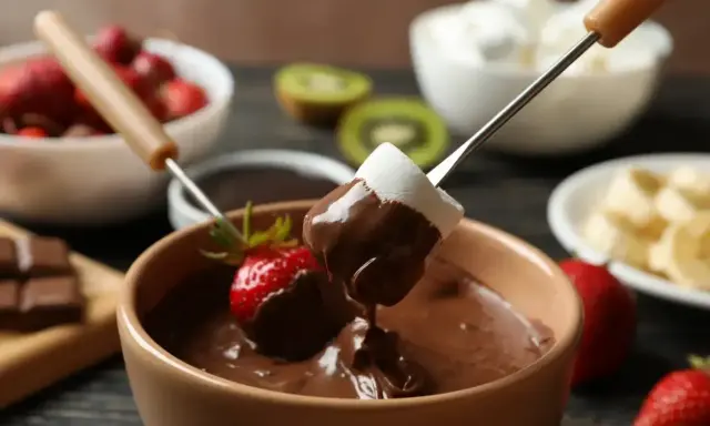 Pyszne fondue czekoladowe z pianką i truskawką na patyczku. W tle owoce i pianki.