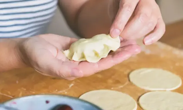 Dłonie formują pieroga z ciasta na pierogi bez jajka. Na blacie leżą okrągłe placki ciasta i miska z wiśniami.