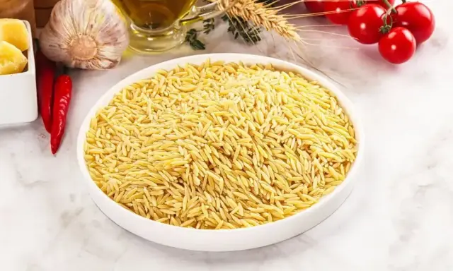 Biała miska pełna makaronu w kształcie ryżu, obok czosnek, chili, oliwa i pomidorki.
