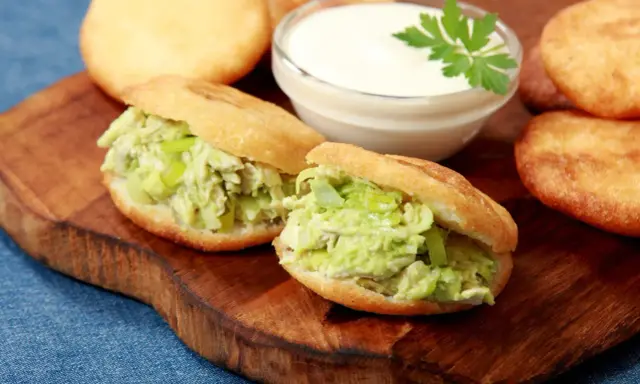 Dwie pyszne arepas z kurczakiem i awokado, obok miseczka sosu i kilka niefaszerowanych arepas.