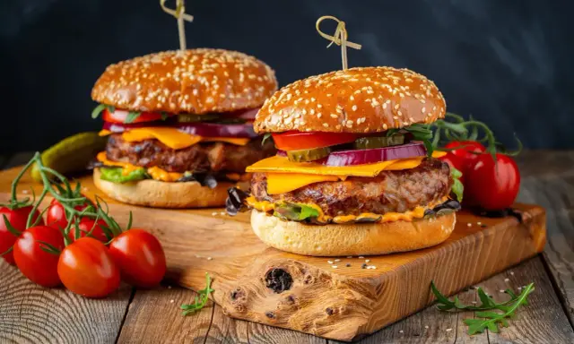 Dwa soczyste burgery z idealnie wysmażonym mięsem na burgery, serem, warzywami i sezamową bułką, podane na drewnianej desce z pomidorkami.