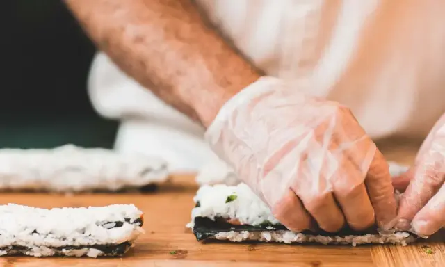 Ręce w rękawiczkach zwijają sushi. Na desce leżą gotowe rolki i składniki do kolejnej. Idealna zalewa do sushi podkreśli smak.