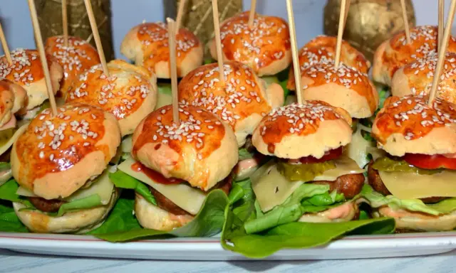 Mini hamburgery z sezamem na patyczkach, ułożone na talerzu z sałatą i dodatkami.