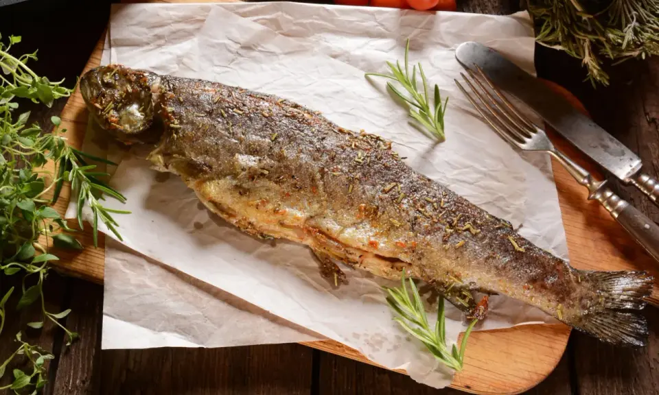 Złocisty pstrąg z patelni, przyprawiony ziołami, leży na papierze do pieczenia. Obok widelec i nóż.