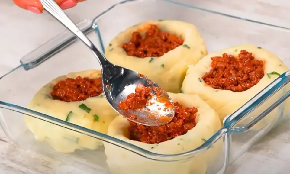 Łyżka nakłada mięsne nadzienie do wgłębień w ziemniaczanych grule, przygotowywanych w szklanym naczyniu.