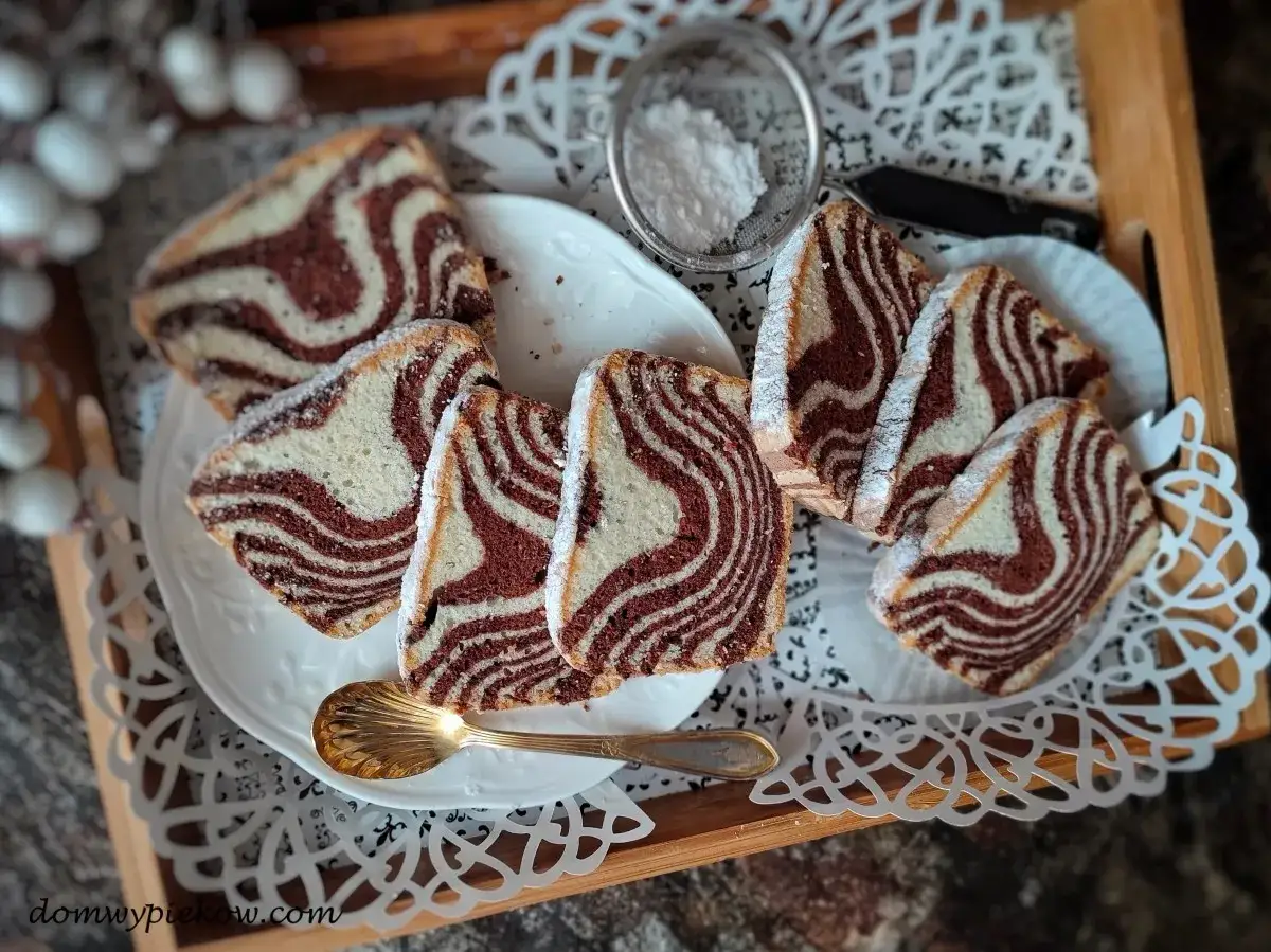 Puszyste ciasto zebra przepis. Pokrojone w plastry ciasto z czekoladowymi i waniliowymi pasami, posypane cukrem pudrem, na białym talerzu.