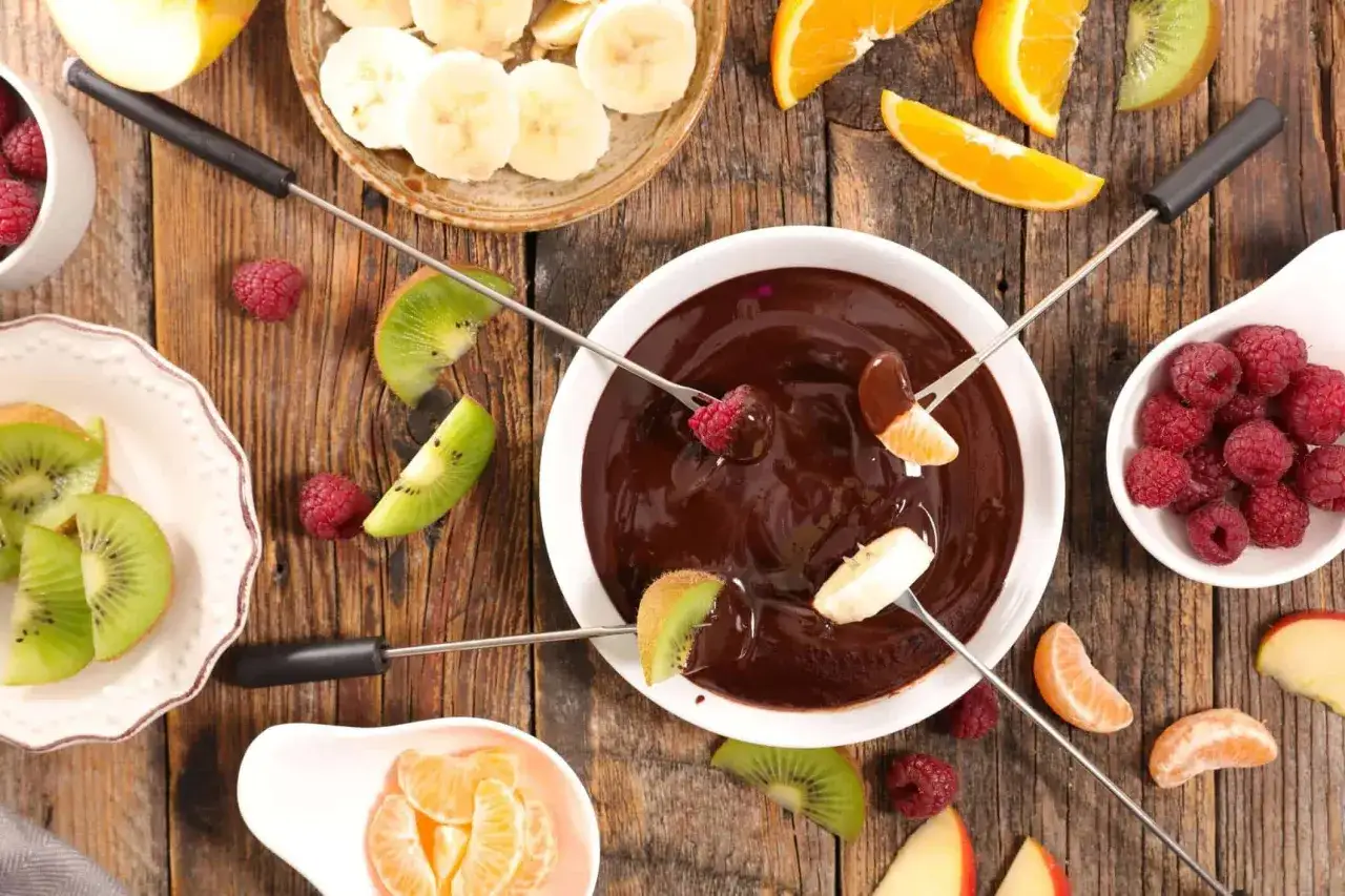 Pyszne fondue czekoladowe z malinami, bananami, kiwi i mandarynkami na drewnianym stole. Idealne na deser!