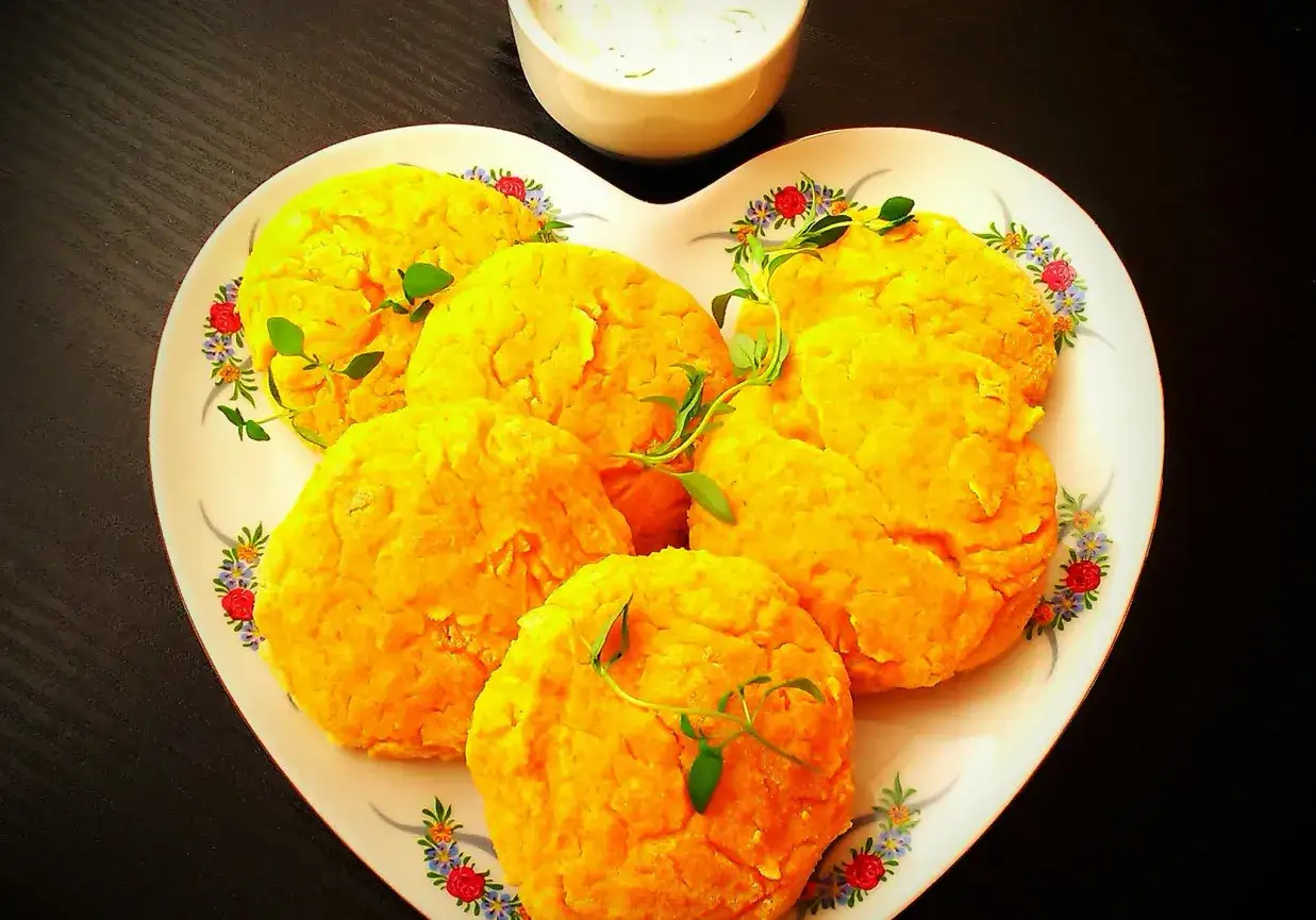Pięć złotych arepas na sercu talerza, ozdobione gałązkami ziół. Obok miseczka z sosem.