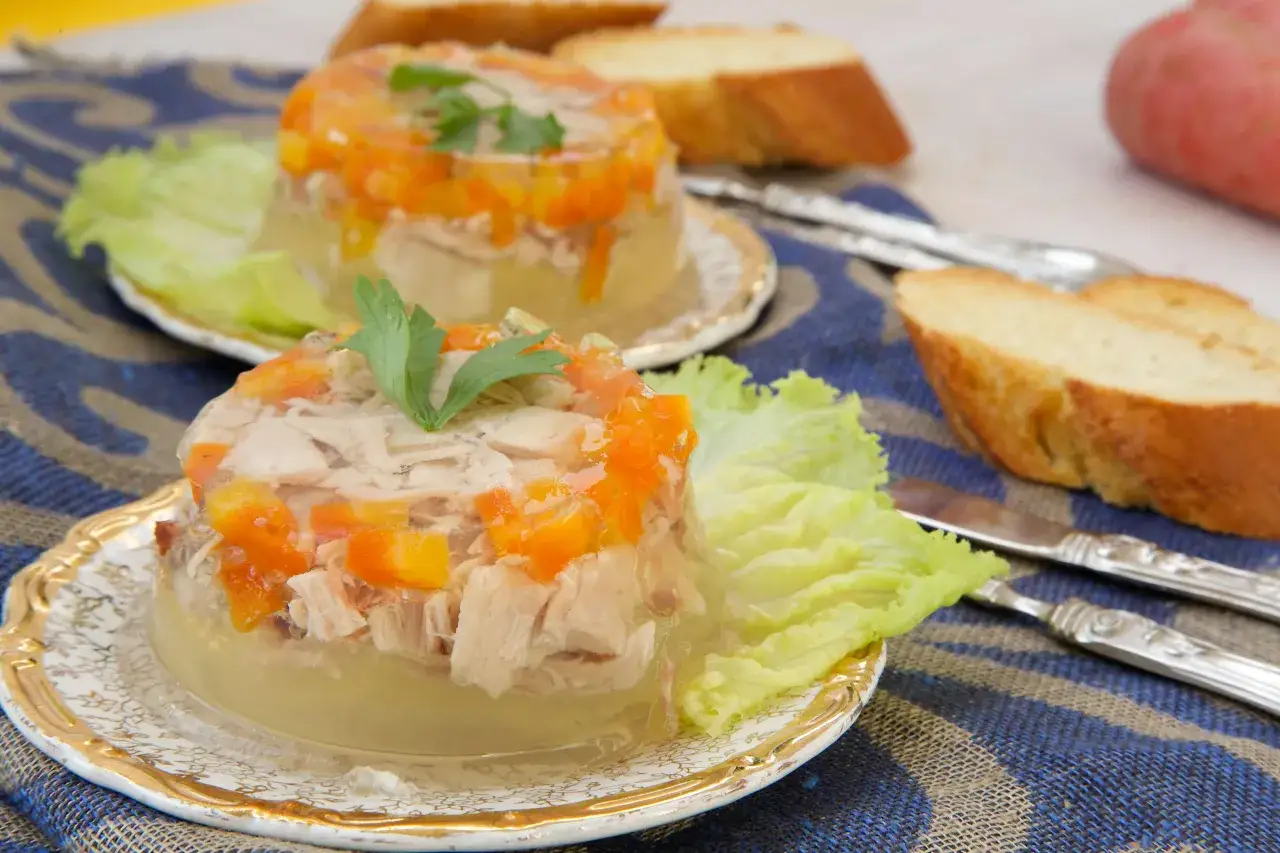 Elegancka ryba w galarecie z marchewką i natką pietruszki, podana na liściu sałaty, obok kromek chleba.