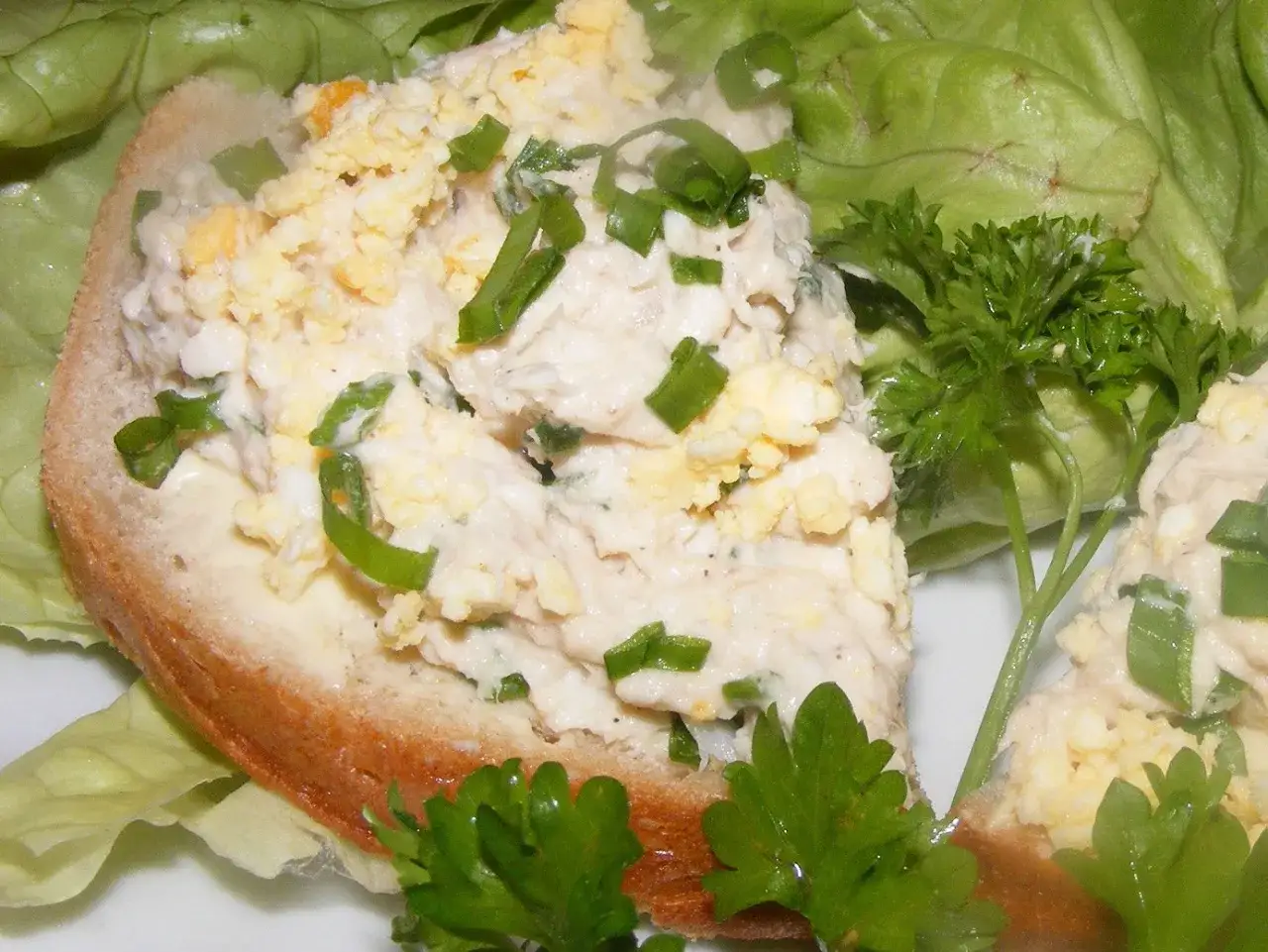 Pyszna pasta jajeczna z makrelą na kromce chleba, ozdobiona szczypiorkiem i natką pietruszki, podana na liściach sałaty.