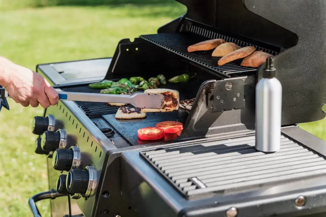 Ręka szczypcami obraca na grillu rumiane plastry sera halloumi, obok pieką się papryczki i pomidory.