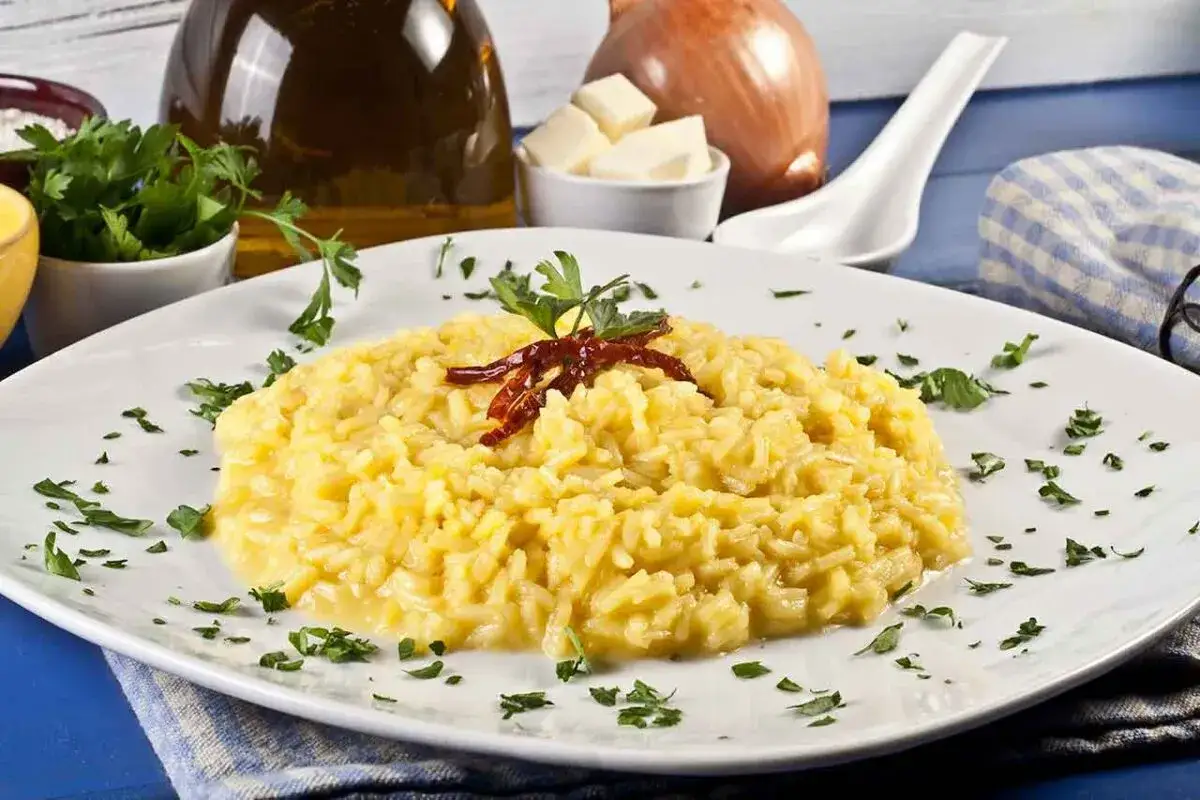 Aksamitne risotto alla milanese, ozdobione natką pietruszki i suszonymi papryczkami. Obok butelka oliwy, cebula i masło.
